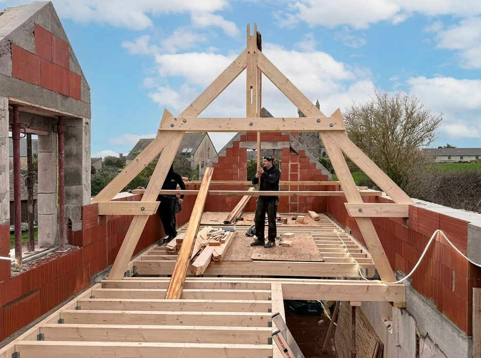 Chantier de charpente traditionnelle en cours, deux charpentiers posent une ferme en bois massif sur des murs en briques monomur