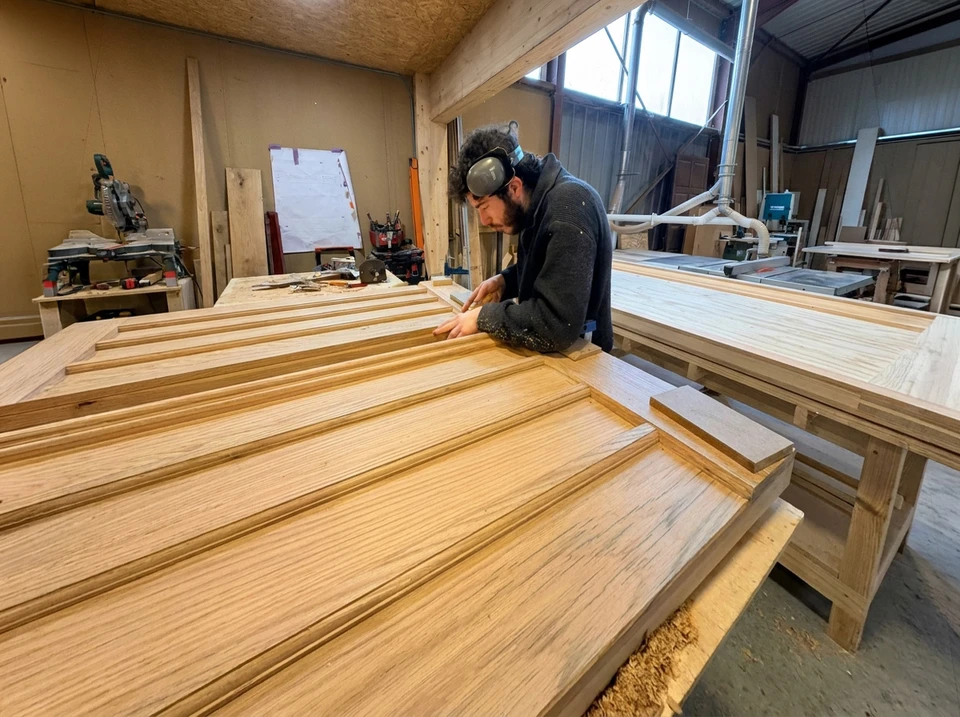 Artisan menuisier travaillant dans son atelier, mesurant avec précision une porte en bois massif à l'aide d'une équerre pliante métallique et marquant au crayon rouge